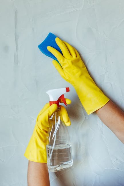 A person wearing yellow rubber cleaning gloves is using a blue sponge to clean a white wall or surface. One hand holds the sponge, while the other grips a clear spray bottle filled with cleaning solut